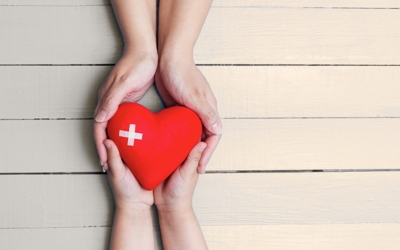 Hände halten ein rotes Herz mit Kreuz.